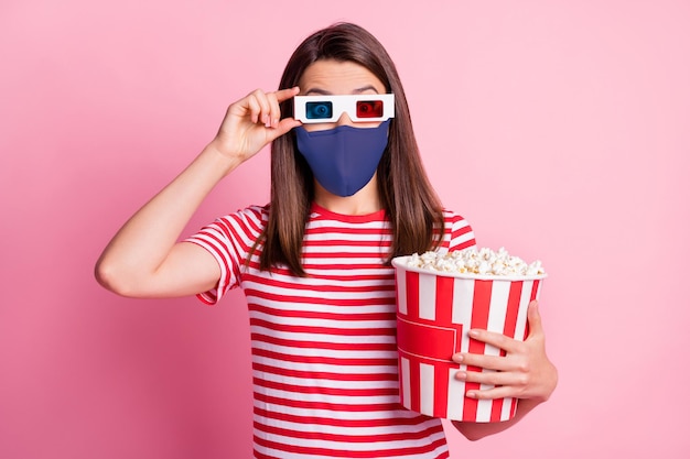 Photo portrait of girl touching 3d glasses holding box with pop corn watching movie wearing blue fabric face mask isolated on pastel pink colored background