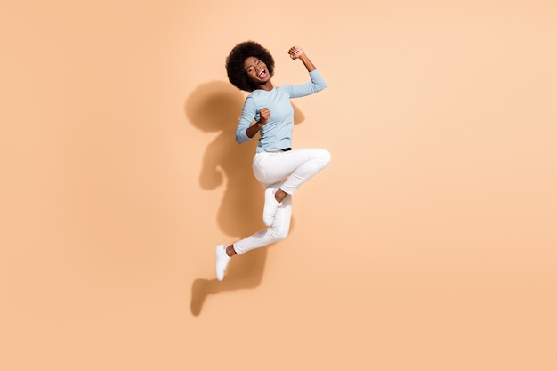 Photo portrait of glad african american woman jumping up holding fists up laughing isolated on pastel beige colored background