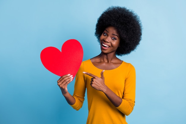 Photo portrait of happy smiling black skinned girl pointing at symbol of valentines day heart card isolated on bright blue color background