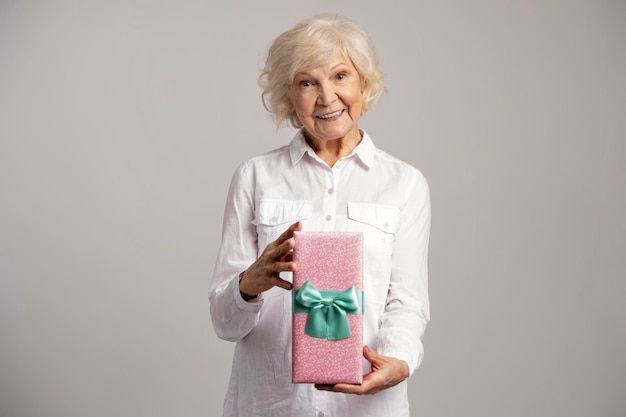 写真 プレゼント ボックスを与え、灰色の背景に分離された笑顔の好奇心旺盛な老婦人の写真の肖像画女性の日の概念