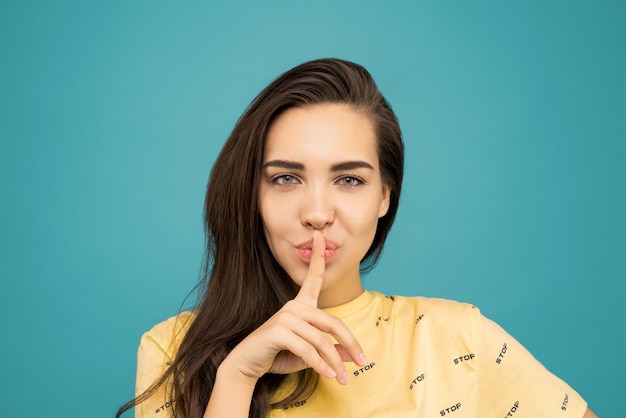 写真 青い背景の前に立っている間shhhサインをしている黄色のtシャツの女性の写真の肖像画