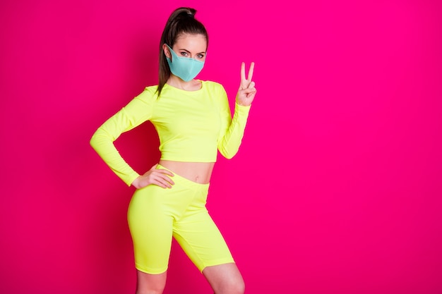 Photo portrait of pretty sportswoman wearing yellow sportswear mask showing v-sign gesture isolated on vivid fuchsia color background