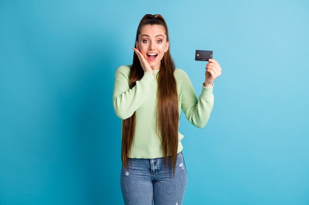 Photo portrait of screaming woman holding plastic card with fingers touching face with hand isolated on pastel blue colored background