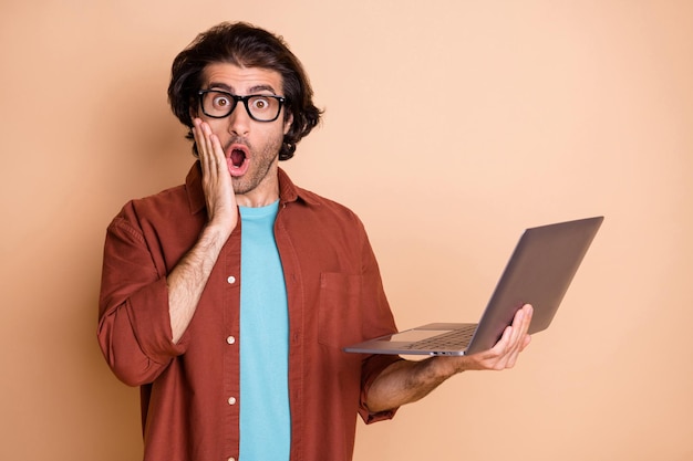 Photo portrait of shocked man touching face cheek holding laptop in hand isolated on pastel beige colored background