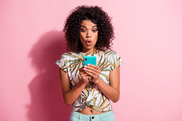 Photo portrait of shocked woman holding phone in two hands isolated on pastel pink colored background