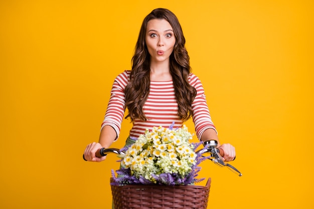 Photo portrait of surprised girl keeping hands on bicycle with basket of wild flowers isolated on bright yellow color background