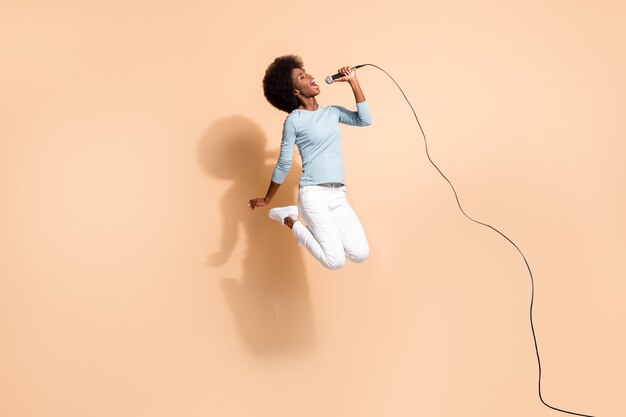 Photo portrait of wild african american woman holding microphone in one hand singing in air isolated on pastel beige colored background