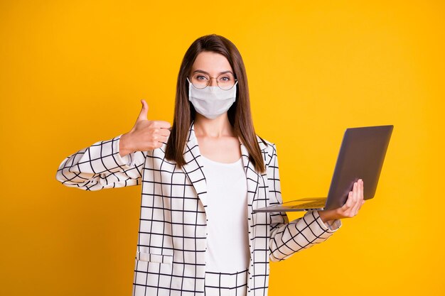 Photo portrait of woman working on laptop showing thumb-up wearing white face mask isolated on vibrant yellow color background