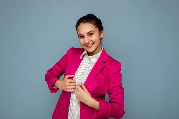 Photo portrait of young attractive beautiful positive smiling brunette woman with sincere emotions wearing casual white shirt and stylish pink jacket isolated over blue background with copy space.