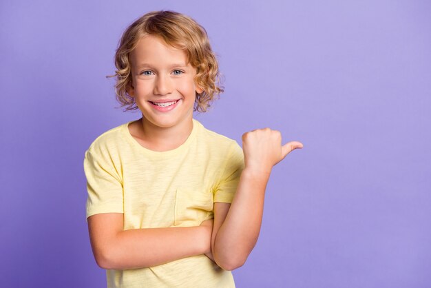 Foto di un ragazzo positivo che indica lo spazio vuoto del dito del pollice per gli annunci promozionali indossa una maglietta gialla isolata su uno sfondo di colore viola