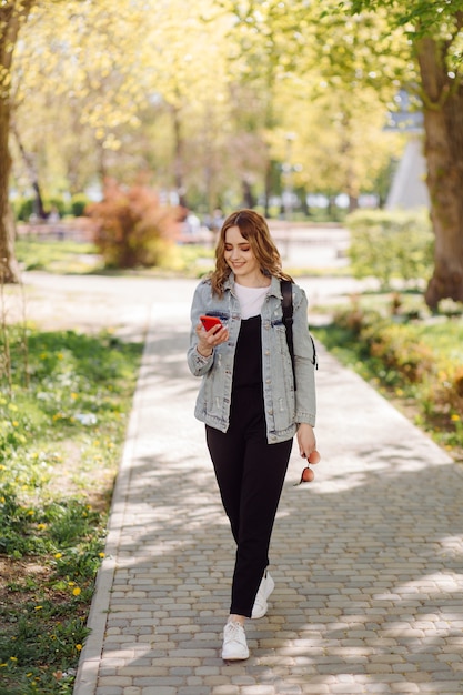 Photo of a positive cheerful teenage girl spends time in the park and using mobile phone.