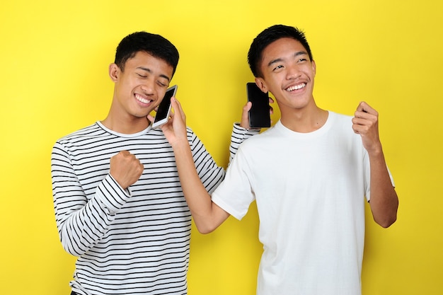 Photo of positive excited two Asian men smiling using mobile phones isolated over yellow background