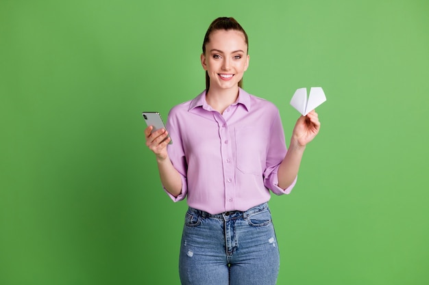 Photo of positive girl hold white paper plane use smartphone send social network mail wear violet lilac shirt denim jeans isolated over green color background