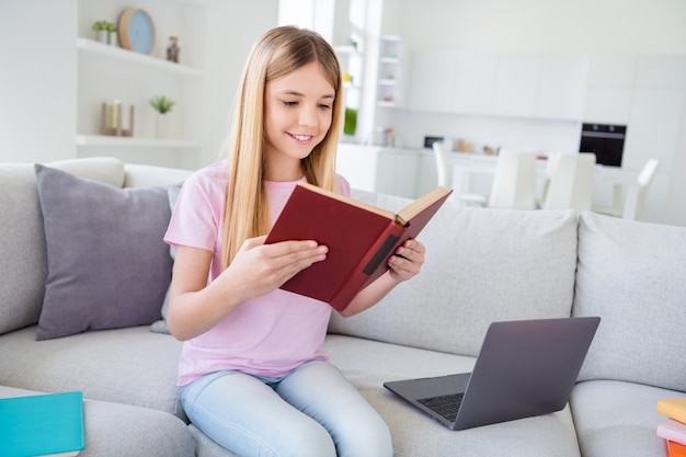 Photo of positive girl kid sit divan study remote read text book prepare homework for tomorrow homeschool lesson wear t-shirt denim jeans in modern apartment house indoors