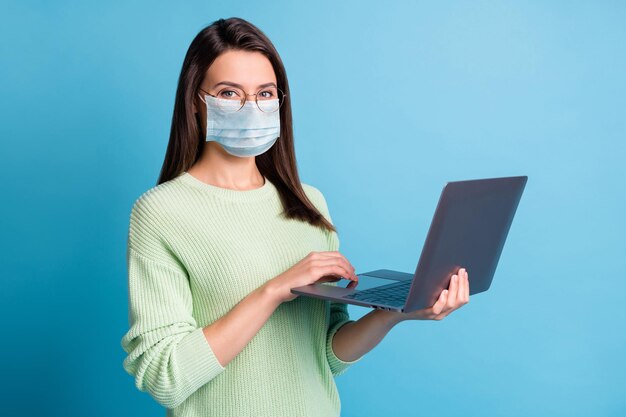 Foto di una ragazza positiva che lavora al laptop indossa una maschera facciale blu isolata su uno sfondo di colore blu