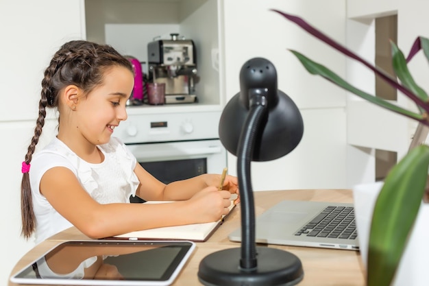 ポジティブな子供の女の子の研究リモートシットデスクテーブル使用ラップトップの写真は、家庭教師が屋内でレポートを書くことでオンラインコミュニケーションレッスンを持っています
