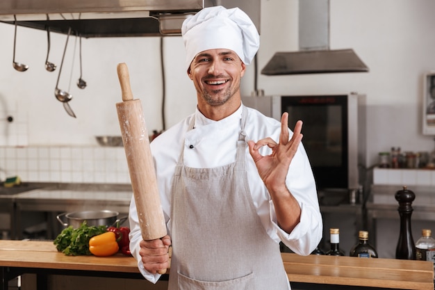 Photo of positive male chief in white uniform holding rolling pin, while standing at kitchen in restaurant