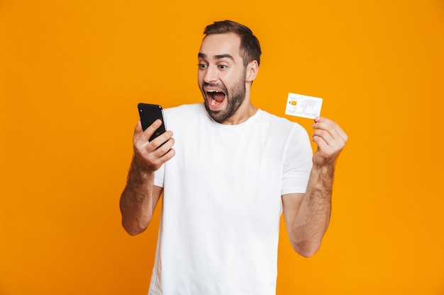 Photo of positive man 30s in casual wear holding smartphone and credit card, isolated