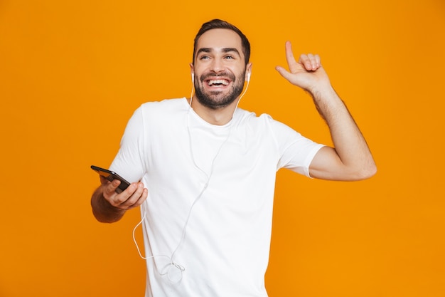 Photo of positive man 30s listening to music using earphones and mobile phone, isolated