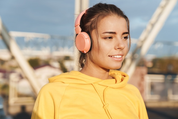 Photo of a positive optimistic pretty young fitness woman posing outdoors listening music with headphones.