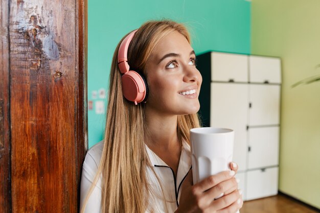 コーヒーを飲みながらヘッドフォンで音楽を聴いて自宅でパジャマを着て前向きな楽観的な笑顔の若いかわいい女の子の写真。