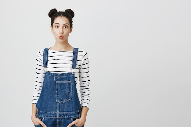 Photo of positive pleasant looking pleased student girl with dark hair in hairbuns poses against studio wall with hands in pockets, wears denim clothes, pouts her lips while thinking about something