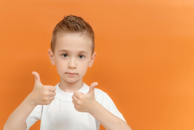 Фотография позитивного дошкольника показывает большой палец вверх знак носить с двумя руками, изолированными над оранжевым цветом
