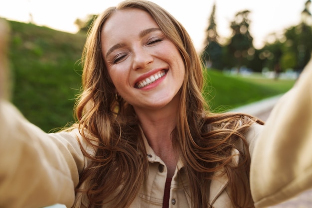 Foto la foto di una bella giovane donna allo zenzero sorridente positiva che cammina nel parco verde della natura all'aperto prende selfie dalla macchina fotografica.