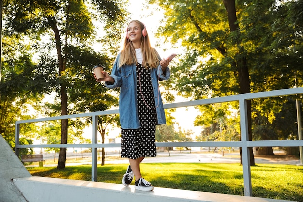 Photo of a positive teenage girl outside in nature green park listening music with headphones using mobile phone drinking coffee.