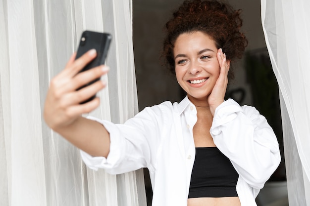 Фотография позитивной молодой женщины, одетой в белую рубашку в помещении в домашнем отеле возле занавески в наушниках, разговаривает по мобильному телефону.