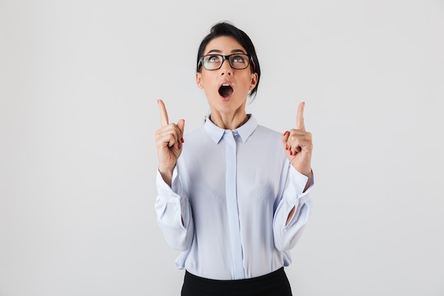 Photo of pretty businesslike woman wearing eyeglasses pointing fingers upward while standing in the office, isolated over white wall