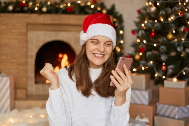 Photo of pretty cute lady clenching fist and having excited facial expression, holds telephone, wears santa x-mas headwear and white sweater, posing at home with x-mas tree and fireplace.