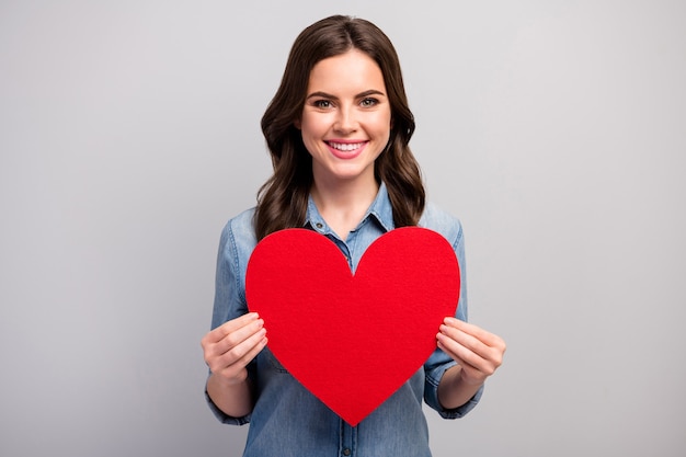 La foto della signora abbastanza divertente tiene il grande cuore di carta rosso che celebra il giorno degli amanti che fa un invito creativo per il colore grigio isolato della camicia del denim dei jeans di usura del ragazzo