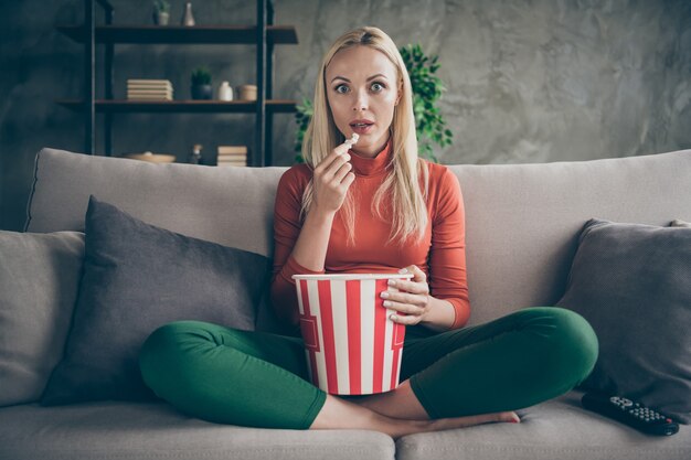 Photo of pretty funny lady homey mood eating popcorn watching television horror show eyes full of fear sitting comfort couch casual outfit flat living room indoors