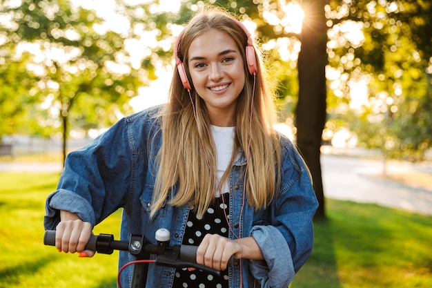Photo of a pretty happy positive optimistic young teenage girl outside in nature green park on grass listening music with headphones walking with scooter.