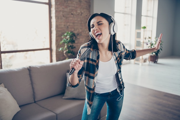 Foto di bella signora che si rallegra ascoltando la melodia preferita nei moderni paraorecchie ballando e cantando nella spazzola per capelli preparazione concerto indossare abiti casual appartamento al chiuso