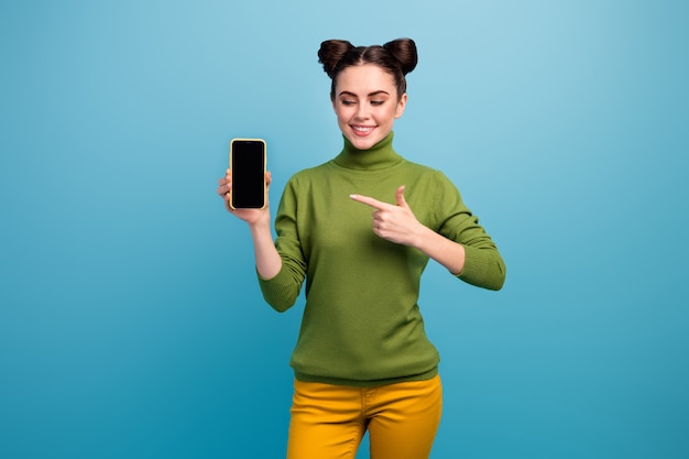 Photo of pretty lady sales manager hold new model smart phone show present advert amazing quality device wear green turtleneck yellow pants isolated blue color wall