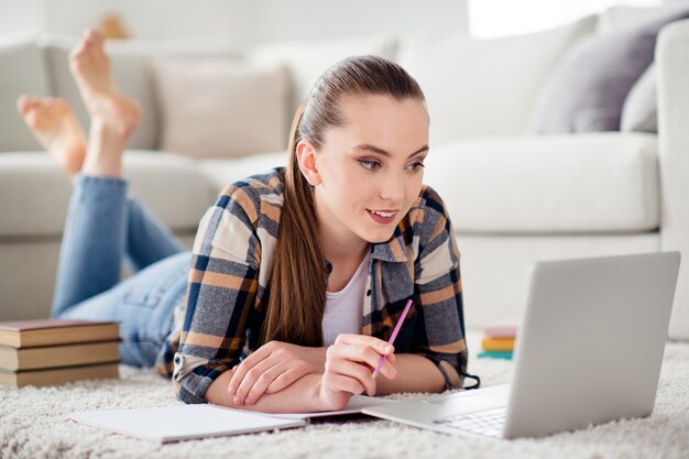 Foto foto di una bella studentessa che sfoglia il quaderno sdraiato sul pavimento guarda la lezione ascolta l'insegnante che scrive appunti