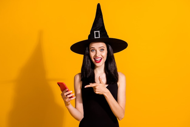 Photo of pretty lovely young magician lady hold telephone directing finger screen smile find curious role play idea wear black wizard headwear dress isolated bright yellow color background