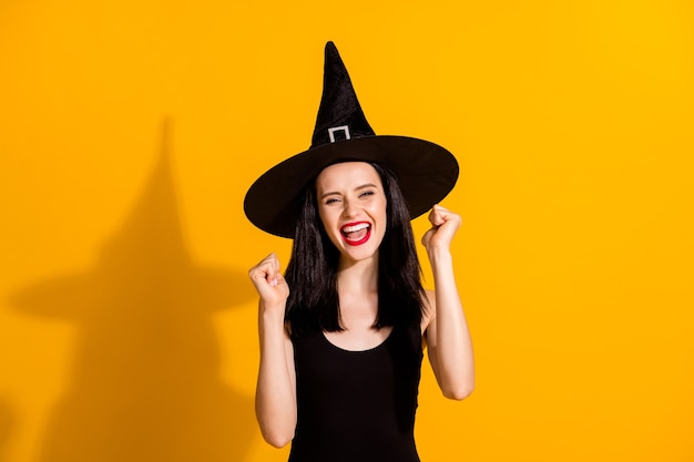 Photo of pretty lovely young magician lady raise fists scream yeah win masquerade quiz excited achieve rare magic item wear black headwear dress isolated bright yellow color background