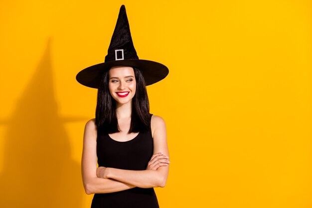 Photo of pretty lovely young sorceress lady folded hands self-assured look empty space sly smile prepare tricks wear black headwear dress isolated bright yellow color background