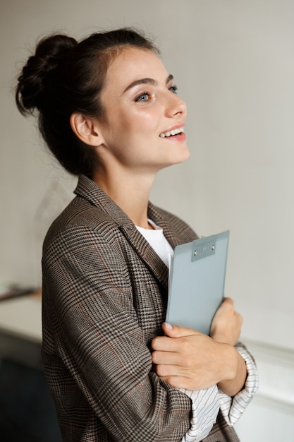 脇を見てクリップボードを保持している自宅で屋内でかなり楽観的な笑顔の若いビジネス女性の写真。