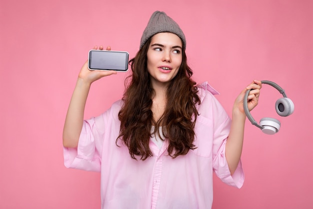 Foto di una giovane donna riccia bruna piuttosto positiva che indossa una camicia rosa e un cappello grigio isolato sopra