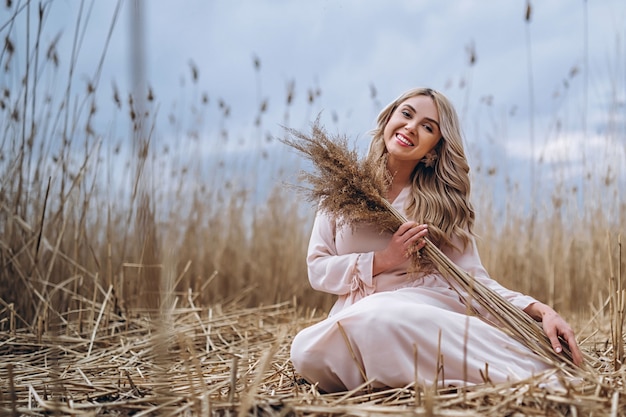 Photo of a pretty smiling girl with long blond curly hair in light long drees sitting in a reed field
