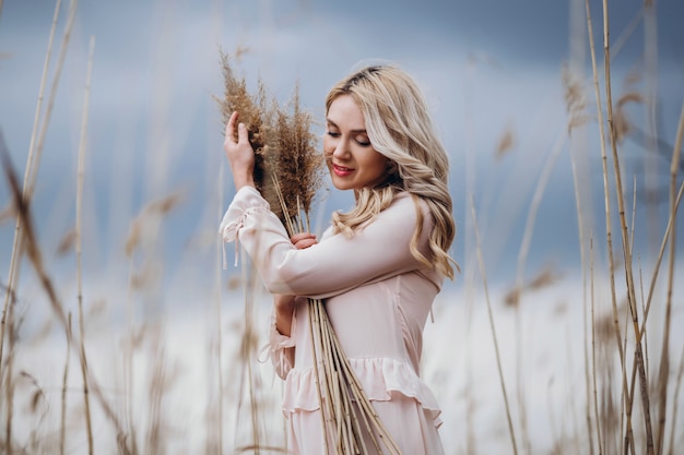 Photo of a pretty smiling girl with long blond curly hair in light long drees standing in a reed field