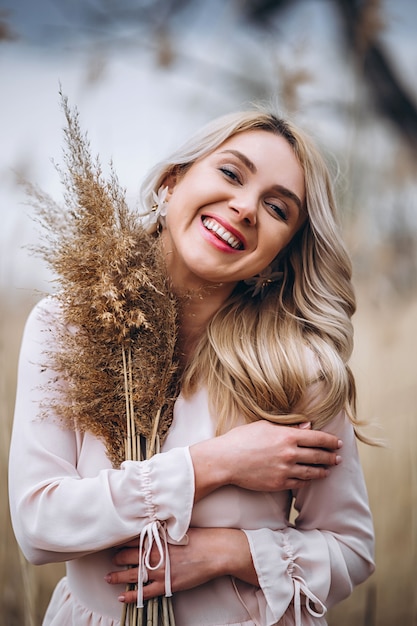 Photo of a pretty smiling girl with long blond curly hair in light long drees standing in a reed field