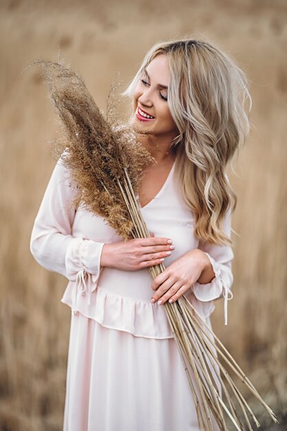 Photo of a pretty smiling girl with long blond curly hair in light long drees standing in a reed field