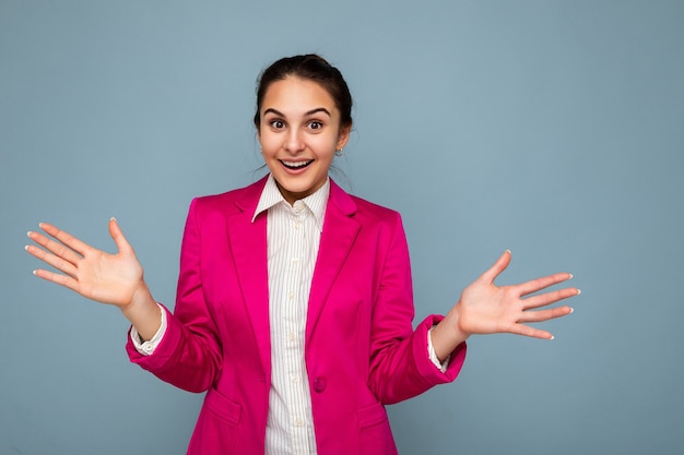 Photo of pretty surprised young brunette woman wearing pink jacket and white shirt isolated over blue background wall looking at camera showing two open palms.