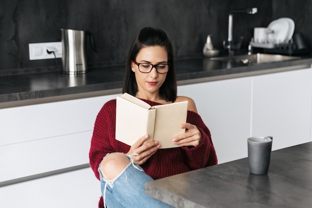 Фотография красивой женщины в помещении дома на кухне, читая книгу.