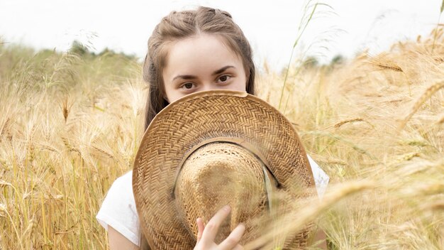Photo of a pretty young girl smiling and covering her face with a straw hat while walking